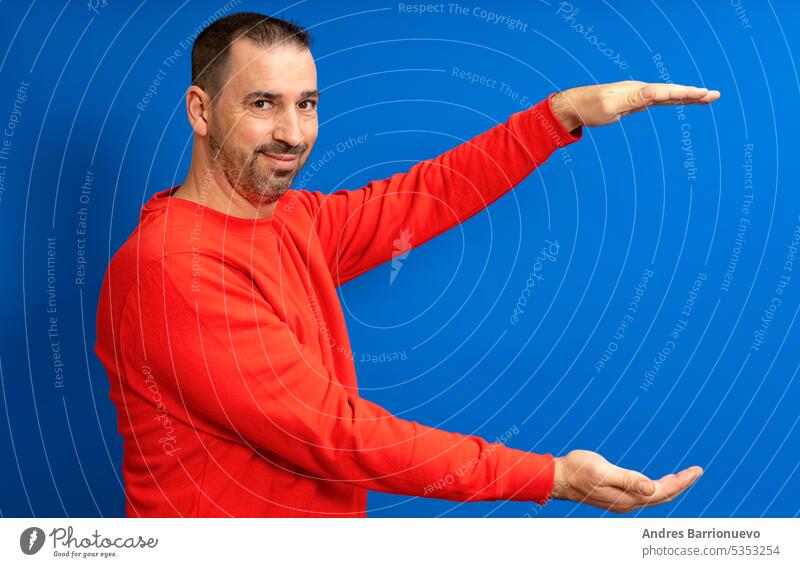 Bearded hispanic man wearing a red sweater with open arms in profile offering something while smiling at camera, isolated over blue background. person handsome