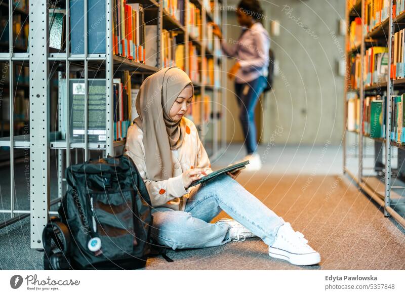 Female muslim student sitting on floor in library and reading book with friend real people teenager campus positive exam knowledge confident academic adult