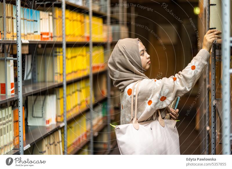 Asian muslim female student picking book from bookshelf in library real people teenager campus positive exam knowledge confident academic adult lifestyle