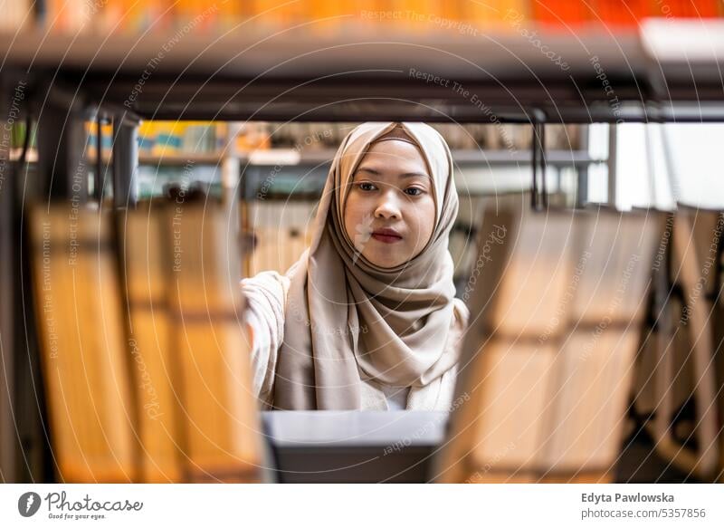Asian muslim female student picking book from bookshelf in library real people teenager campus positive exam knowledge confident academic adult lifestyle