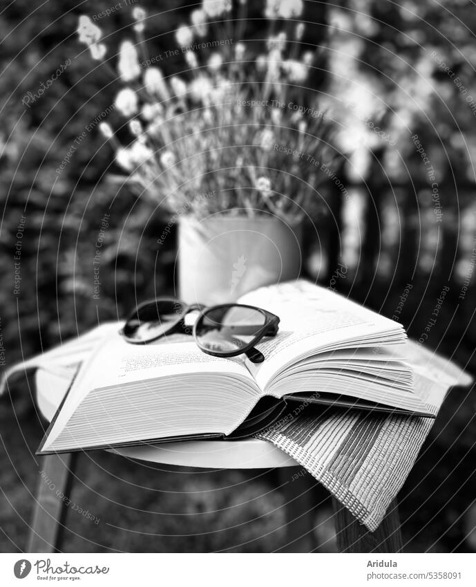 In the garden on a small table is an open book with black glasses. In the background is a white lavender in a pot b/w Eyeglasses Sunglasses Book Reading Garden