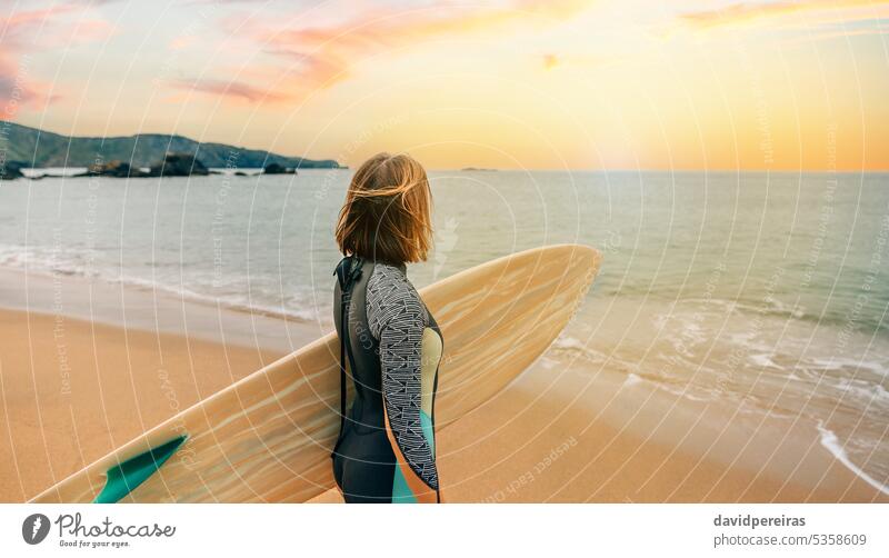 Surfer woman with wetsuit carrying surfboard looking to the sea at the beach unrecognizable young surfer sunset sunlight horizon coast sand summer people 20s