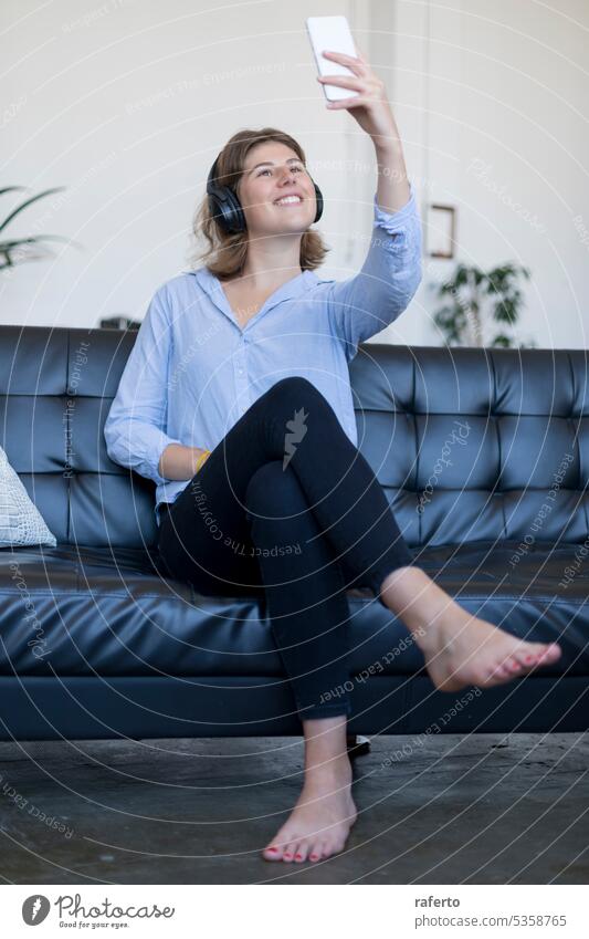 Young smiling woman sitting on leather sofa with headphones taking a selfie young happy joyful girl relaxation technology music leisure lifestyle modern trendy