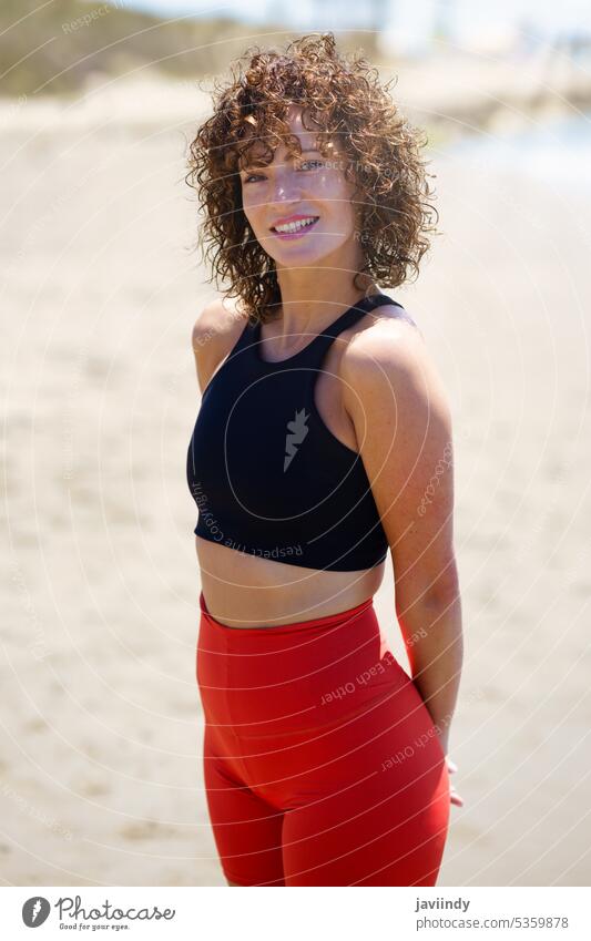 Smiling woman in sportswear standing on beach smile positive athlete fit activewear summer sun hand behind back sea happy curly female leggings fitness
