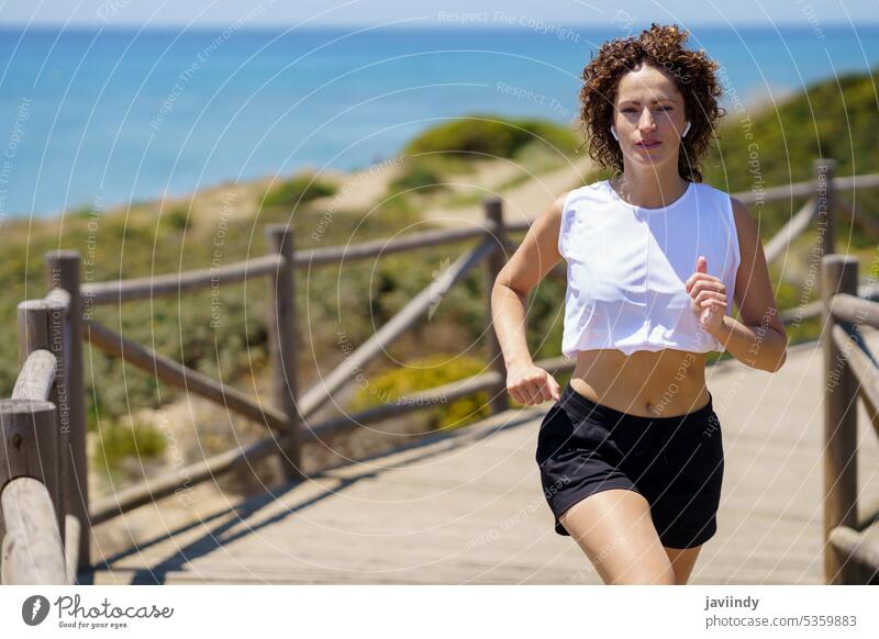 Fit woman jogging on wooden footpath on sunny day sportswoman run workout healthy exercise vitality fit confident beach earbuds listen sea walkway music