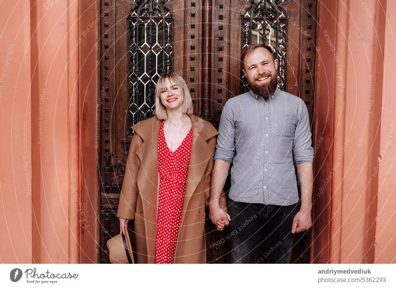 Beautiful stylish happy hipster couple holding sensually hands. Smiling blond woman with short hair and bearded man having romantic date at old European city and looking at camera. Valentines day