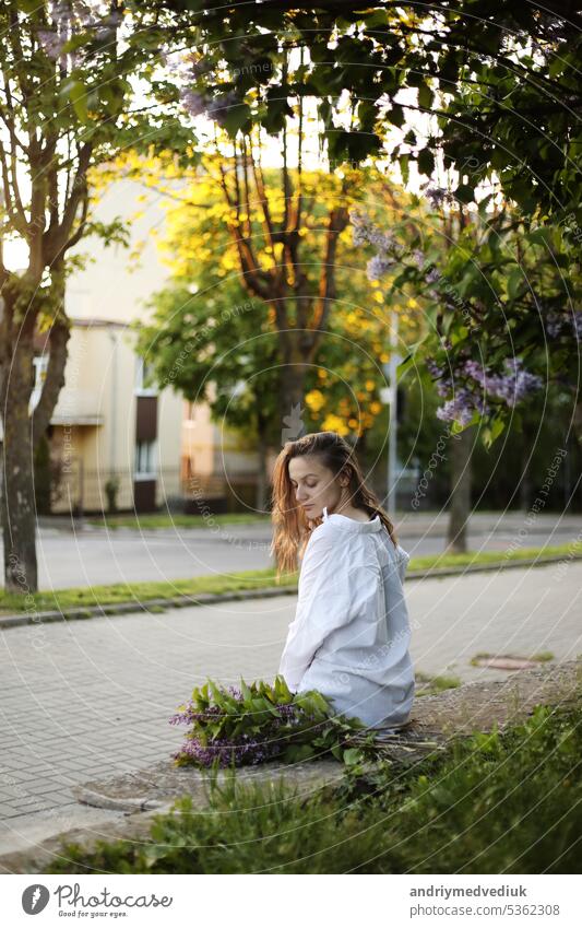 young beautiful girl in a white shirt and black pant sitting near blooming lilac trees with purple flowers outdoors, flowering tree, spring, sun, happiness, tenderness.
