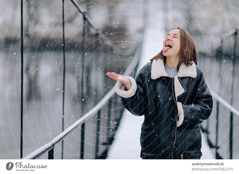 A woman walks and catches snowflakes with his tongue over the river on a suspension bridge in winter day. Young girl in warm clothes stands on a wooden footbridge in cold snowy day