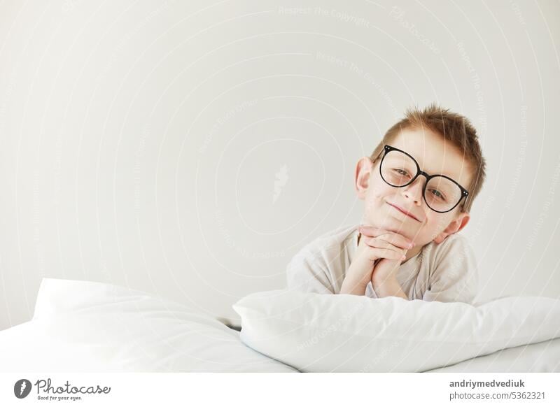 Adorable smiling child boy in glasses is lying in bed. morning dreaming on white bed. childhood sleeping bedroom baby fashion hand love person book kid school