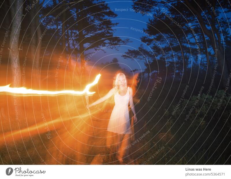 On a dark summer night, a gorgeous blonde girl dressed in a white dress is wandering around with a fire torch. Some long exposure adds suspension and spookiness to the shot. Dark blue sky, green woods, and a pretty woman in this one.
