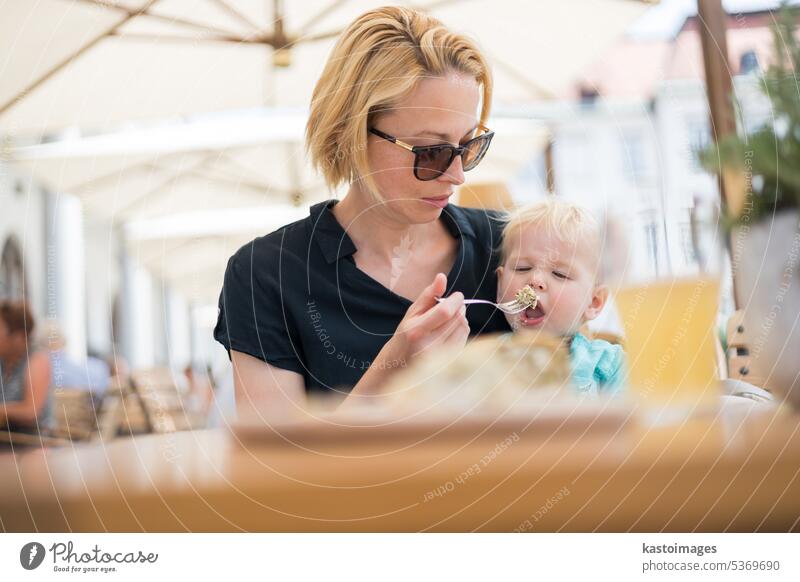 Young caucasian blonde mother spoon feeding her little infant baby boy child outdoors on restaurant or cafe terrace in summer. family together food toddler