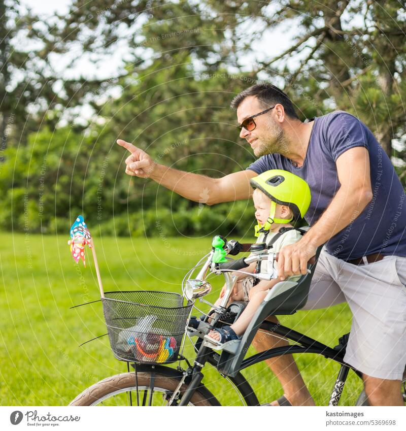 Look over there. Active family day in nature. Father and son ride bike through city park on sunny summer day. A cute boy is sitting in front bicycle chair while father rides bicycle. Father son bonding.