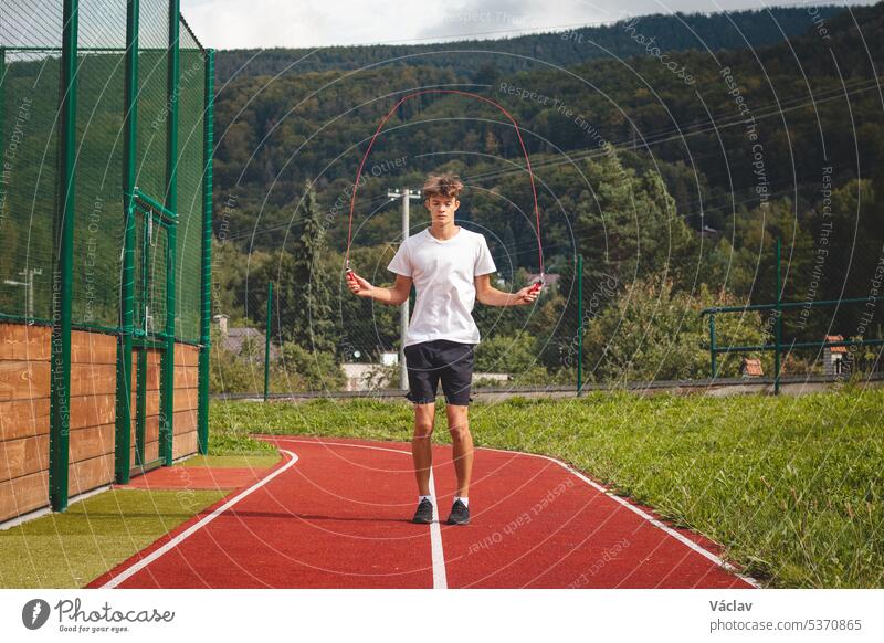 Brown-haired boy with an athletic figure wearing a white T-shirt and black shorts is jumping rope on an athletic oval. Training to improve jumping, coordination and endurance strength