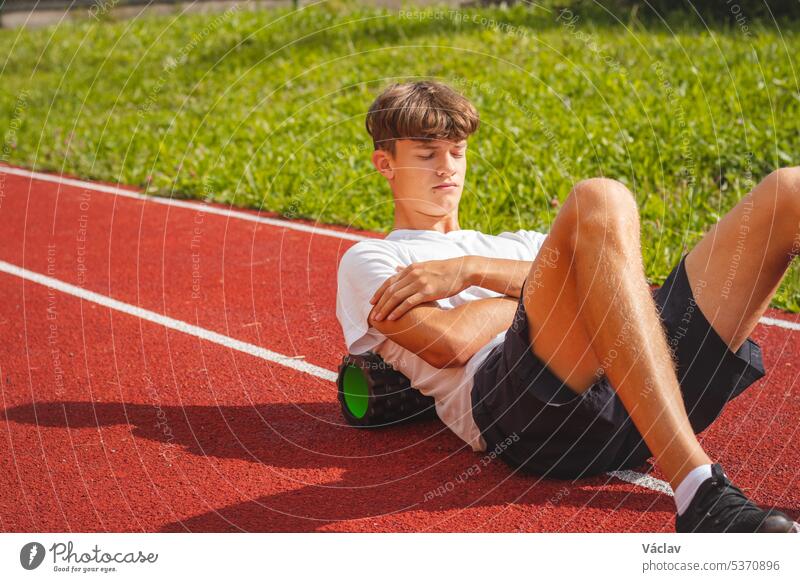 Brown-haired boy with an athletic physique on an athletic oval massages his muscles with a foam roller for better recovery. Post-workout strength abdominal
