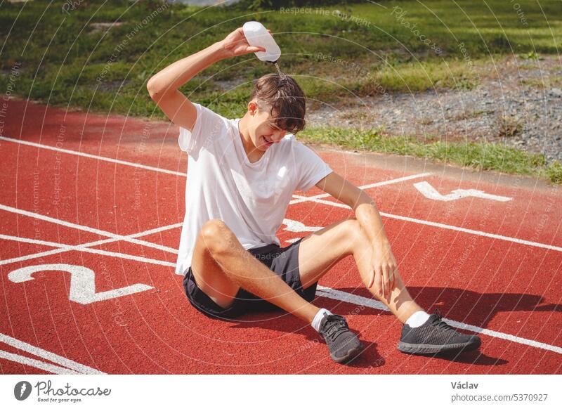 Young athlete refreshes himself with water after a hard workout on the athletic oval in the intense heat. Endurance training. Brown-haired adolescent person
