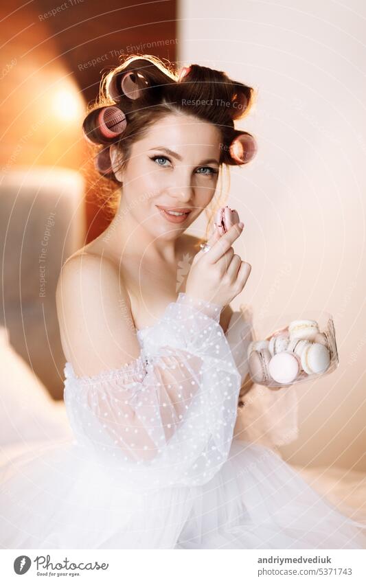 Beauty portrait of a beautiful smiling young brunette woman with bright pin-up make-up eating delicious french macaroons or macarons. girl in white dress and pink curlers sitting on the bed and relax