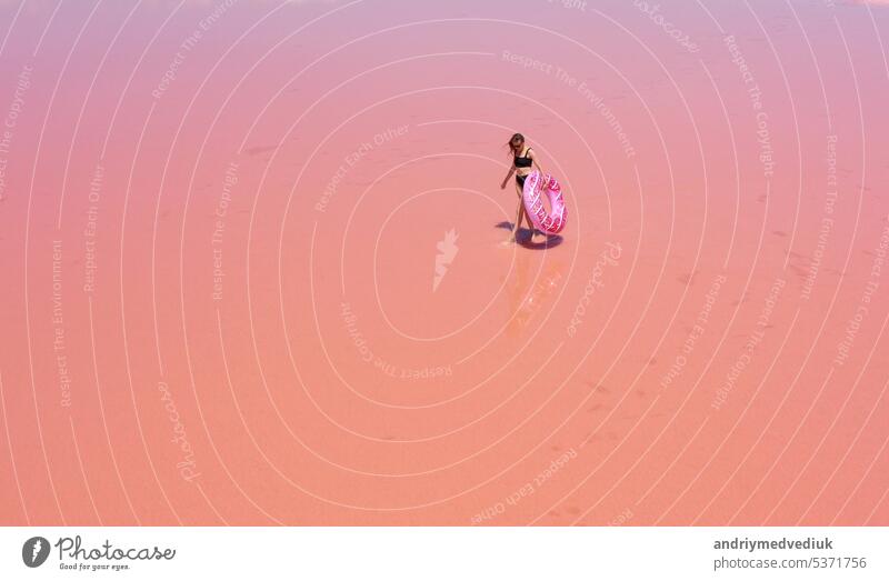 Aerial view. Young woman in black swimsuit carries an inflatable pink float in her hands have fun dancing on the pink salt lake. Concept summer festivals, holiday, travel vacation, freedom, sun, enjoy