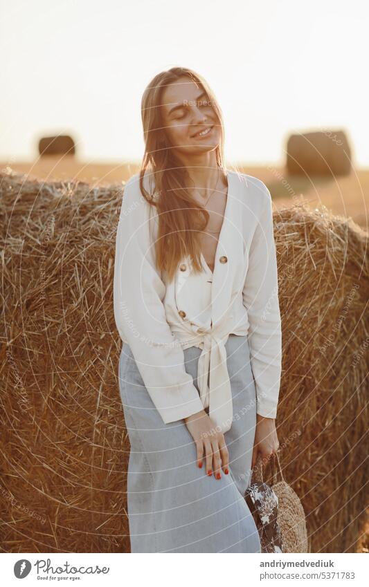 Full length portrait of a smiling beautiful brunette in a jeans skirt and straw bag in hand. Woman enjoying a walk in a wheat field with hay bales on summer sunny day.