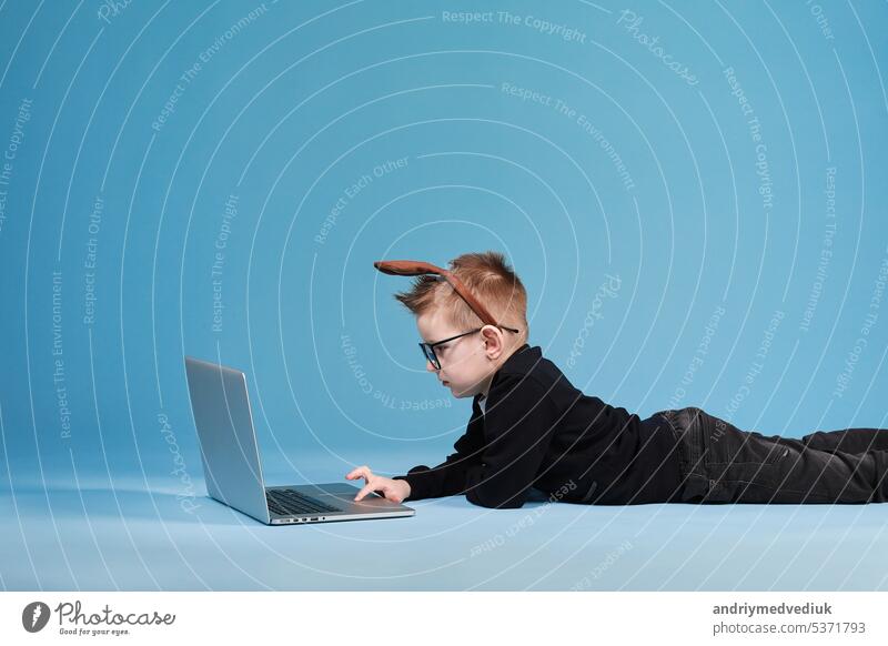 Distance learning online education. Schoolboy in rabbit bunny ears on head studying at home with laptop and doing school homework on blue background kid