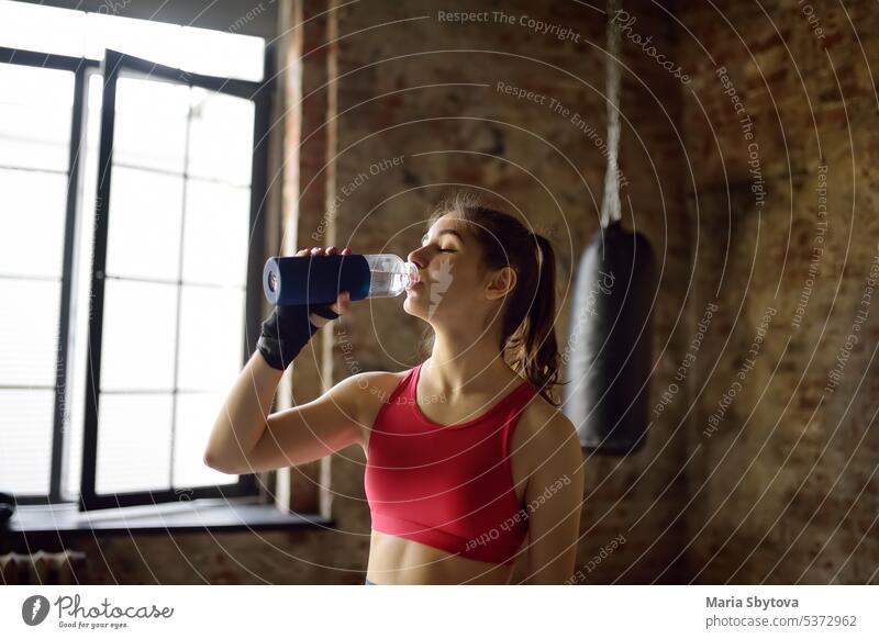 Portrait of young beautiful athlete in a boxing gym. A boxer girl drinking beverage from a bottle during a workout. In the process of training need to drink a lot water.