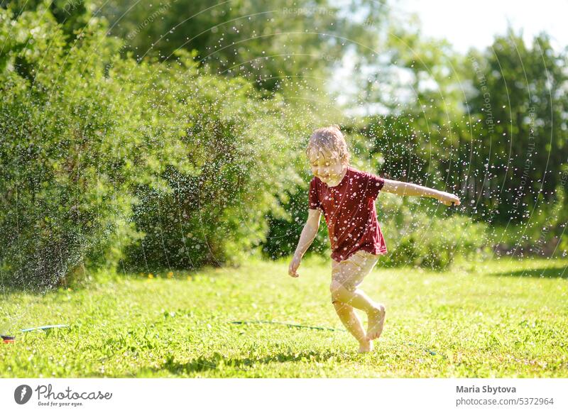Funny little boy playing with garden sprinkler in sunny backyard. Preschooler child laughing, jumping and having fun with spray of water. kid activity summer
