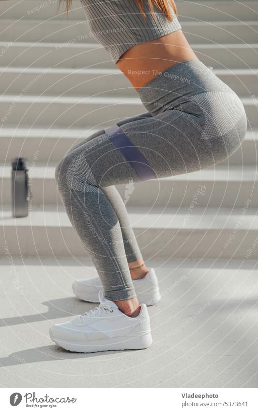 Sporty athletic woman in grey sportswear doing exercises with gymnastic elastic bands sit-ups outdoors workout training body buttocks squat legs tracksuit