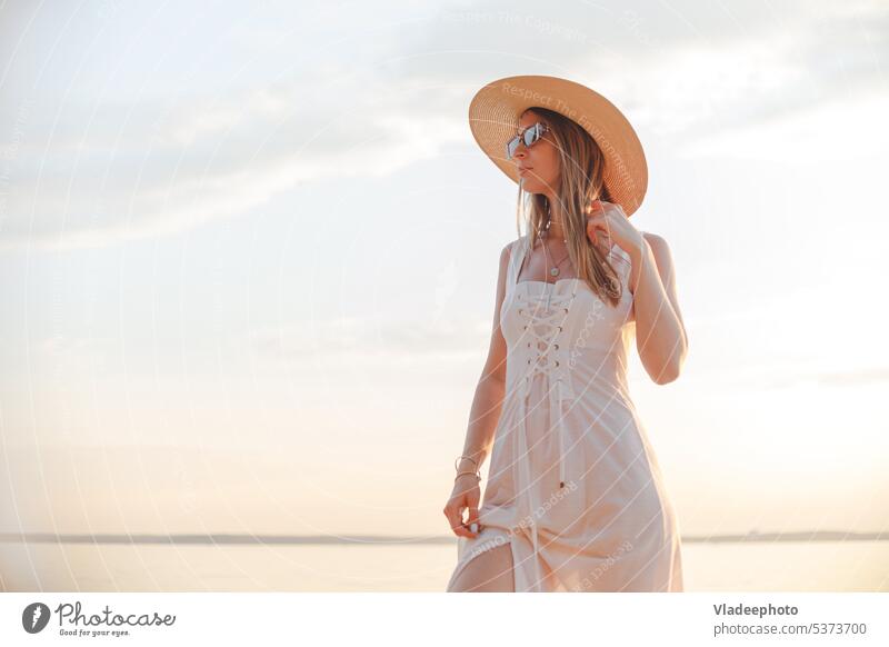 Young caucasian woman in white dress, summer hat and sunglasses posing on beach on sunset sea attractive smiling ship vibe travel beautiful female portrait