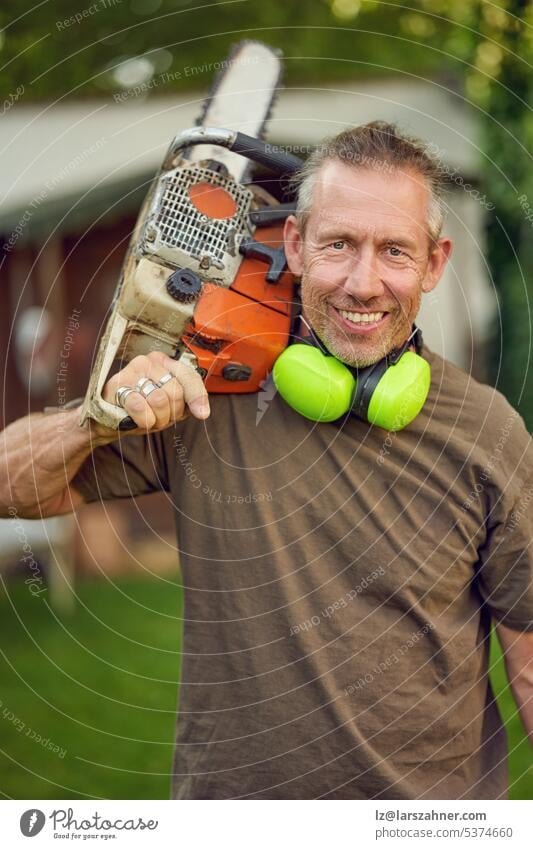 Middle-aged gardener holding a chainsaw over his shoulder after work with ear protection looking at the camera craftsman outside concept craftsmanship worker