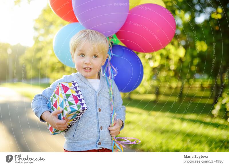 Cute little child going to congratulate a friend on his birthday. Toddler holding bundle of colorful balloons and gift in a festive box. Anticipation of the holiday.