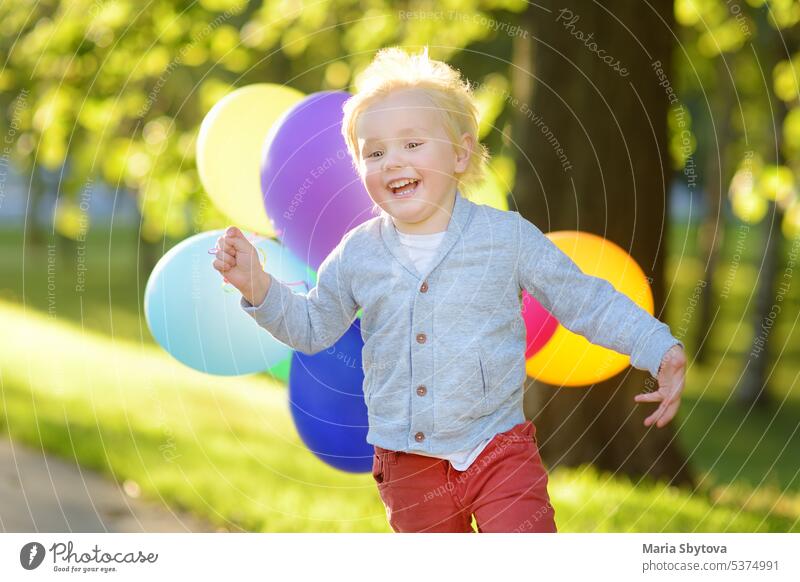 Little boy having fun during celebrating birthday party. Happy child with a bunch of bright multicolor balloons. Preschoolers or toddlers baby birthday party in sunny park. Summer outdoor festival