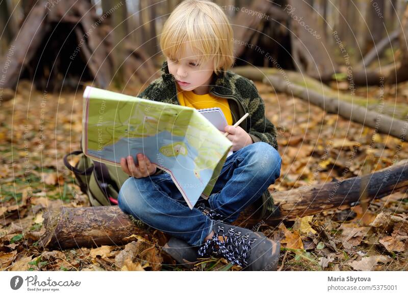 Little boy scout is orienteering in forest. Child is sitting on fallen tree and looking on map on background of teepee hut. child hiking kid navigation