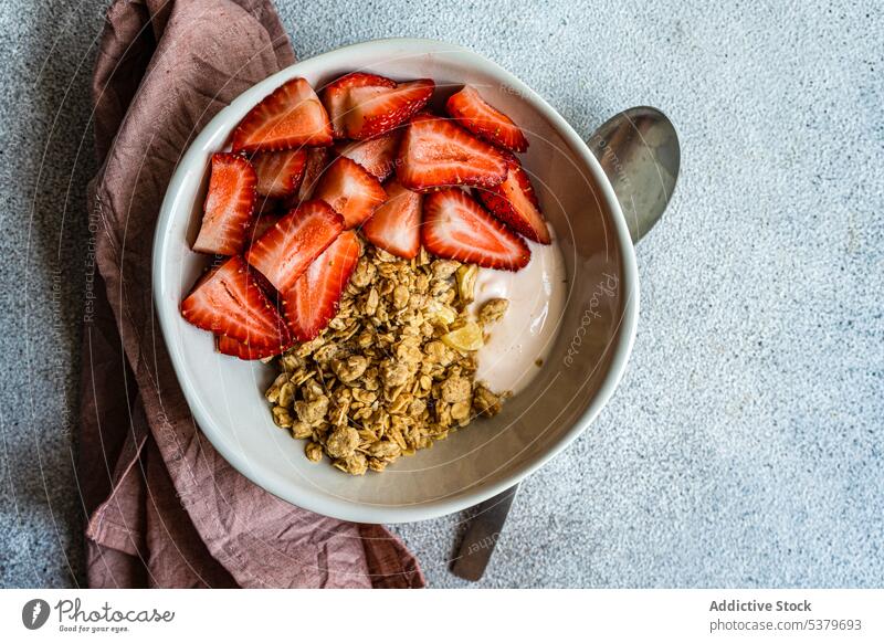 Top view of bowl with granola, yogurt and ripe organic strawberries on grey surface with spoon and napkin background berry breakfast cereal concept concrete