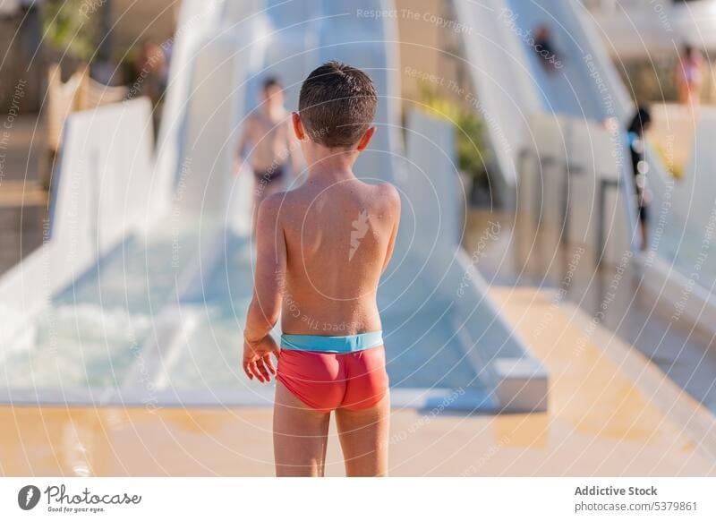 Anonymous kid standing on near slide exit child swimwear shirtless bare shoulders summertime calm childhood vacation boy water weekend recreation playful
