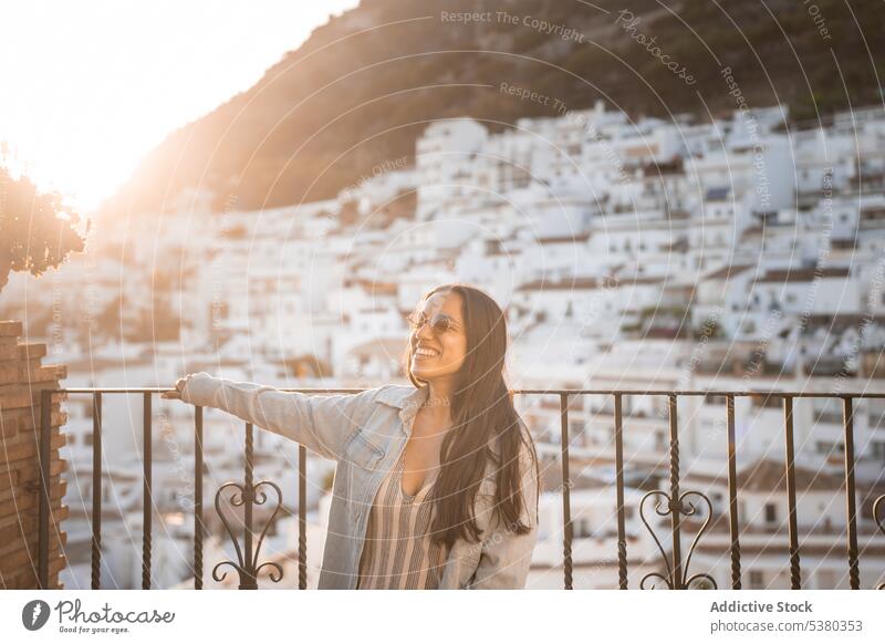 Young woman in sunglasses standing on balcony town smile enjoy tourist mountain vacation fence building young female happy cheerful summer house admire traveler