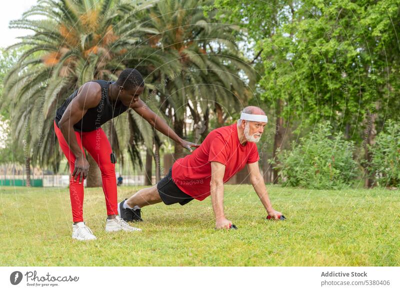 Serious black trainer helping man to do push ups on lawn men training workout exercise park sport athlete functional senior focus wellness male healthy