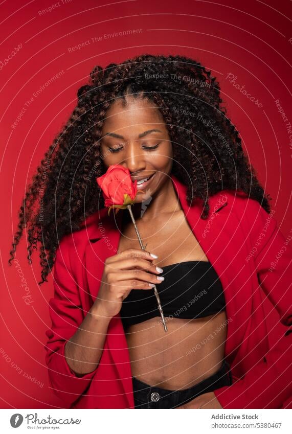 Sensual woman biting rose stalk in red studio romantic flower smile style model happy trendy cheerful young female black eyes closed fashion positive glad
