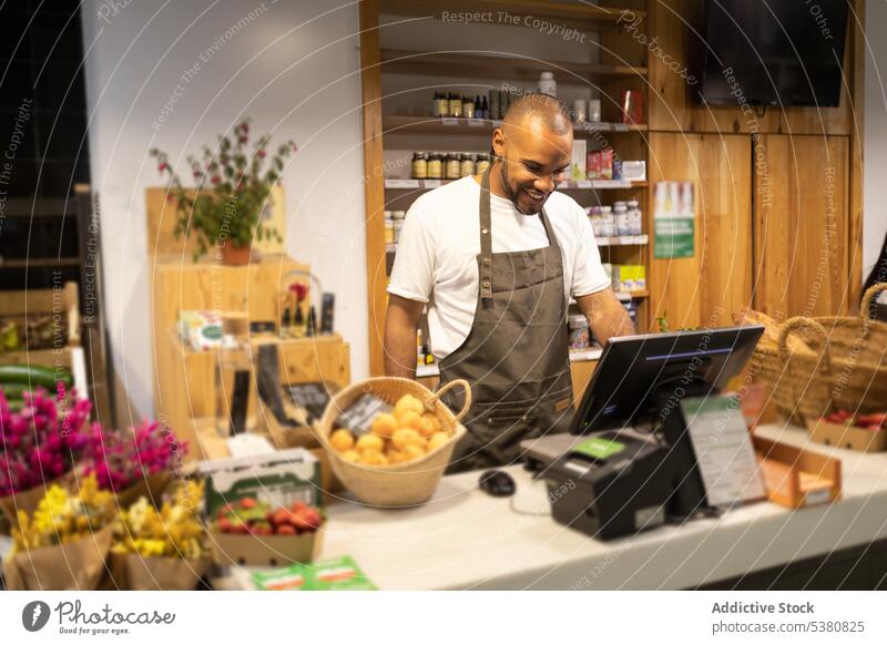 Positive black man using computer during work in store counter shop seller sale register positive cashier supermarket smile staff african american male apron