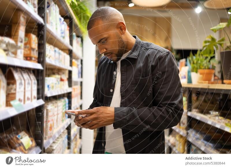 Concentrated black man using smartphone in grocery store shop customer supermarket purchase choose message buy male african american ethnic casual young buyer