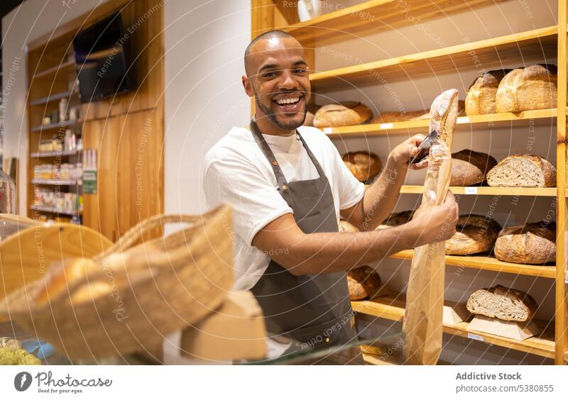 Cheerful black man putting bread in paper bag bakery seller grocery shop cheerful small business work baked owner food staff male pastry job store smile