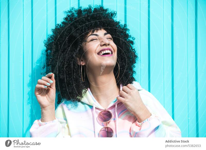 Cheerful ethnic woman laughing on blue background fun fashion cheerful happy appearance hairstyle shadow wall summer young female hispanic outfit accessory afro
