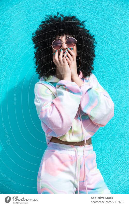 Cheerful ethnic woman standing with hands covering mouth cover mouth laugh fun sunglasses fashion cheerful happy appearance hairstyle shadow wall summer young