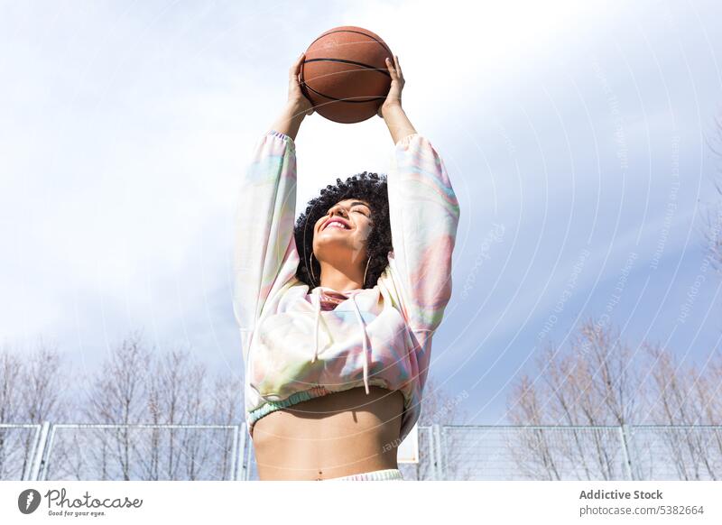 Cheerful ethnic woman holding basketball in stretched hands eyes closed blue sky player smile arm raised court positive summer female young hispanic
