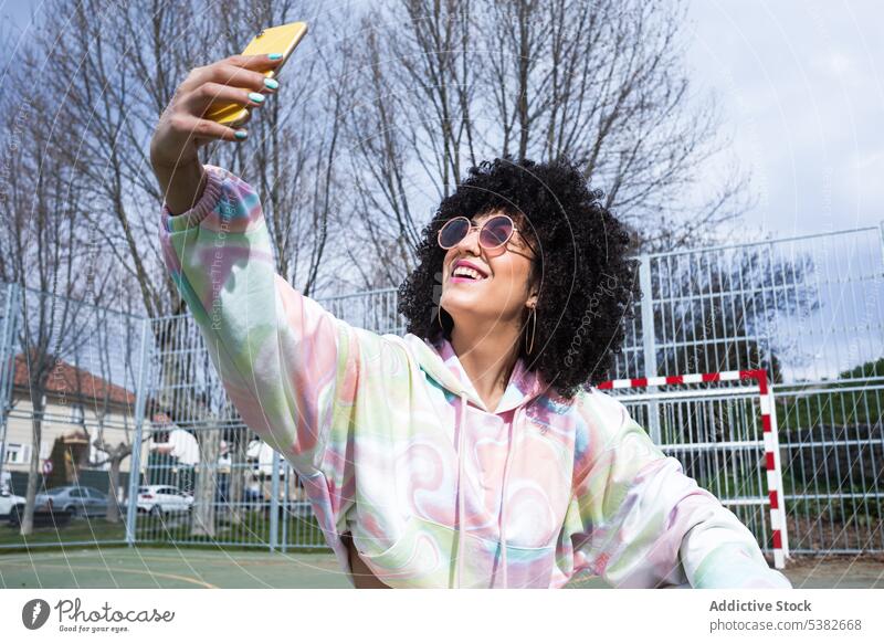 Cheerful ethnic woman taking selfie with smartphone happy cheerful sports ground fence self portrait moment summertime young female hispanic positive sunlight