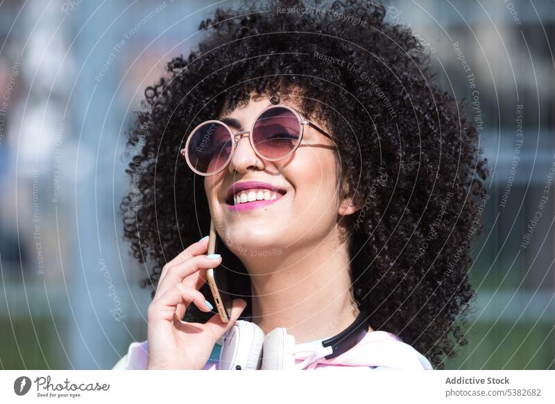 Cheerful ethnic woman standing and speaking on smartphone headphones talk cheerful phone call smile positive conversation young female hispanic street urban