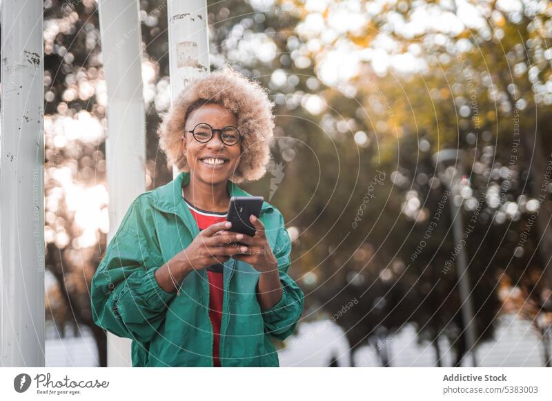 Cheerful young woman using smartphone outdoors cheerful mobile positive browsing smile park female eyeglasses black happy message african american surfing
