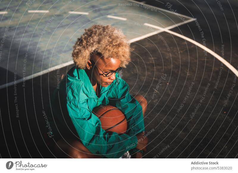 Black woman in curly hair sitting with basketball on ground sportswoman player sports ground evening sportswear rest court young female urban serious jacket