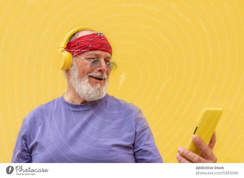 Senior man in headphones using smartphone audio beard entertain song mobile gesture tune move positive happy studio shot listen yellow background male browsing