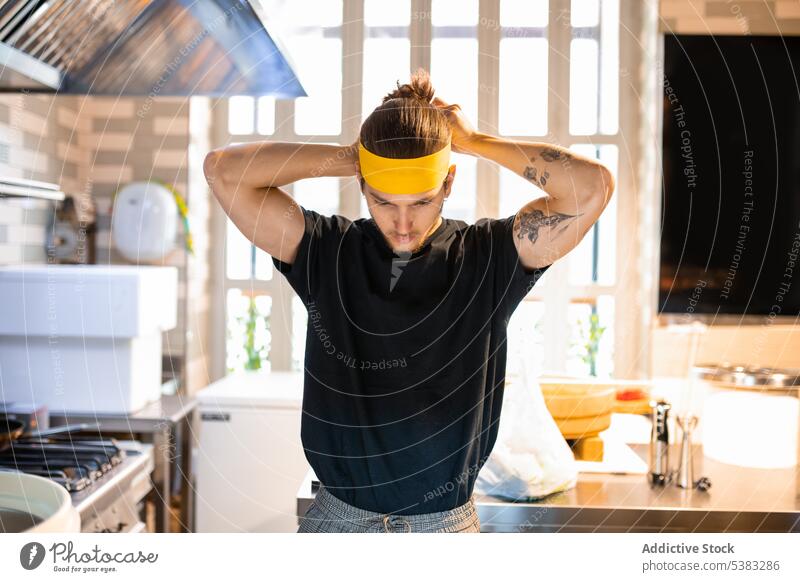 Focused young man tying headband in kitchen before cooking focus chef tie serious concentrate utensil kitchenware male process stove prepare gas culinary