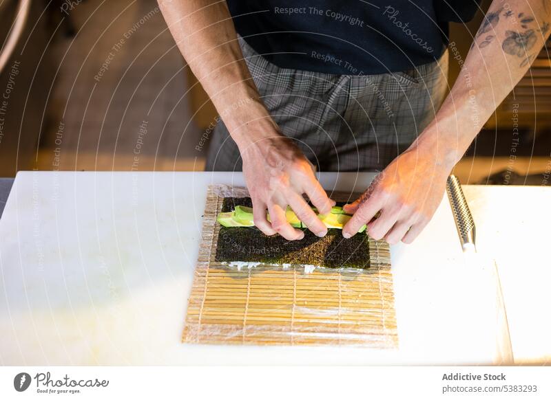 Unrecognizable man arranging avocado slices on seaweed wrap roll anonymous table kitchen prepare ingredient food bamboo male dish wooden organic apron