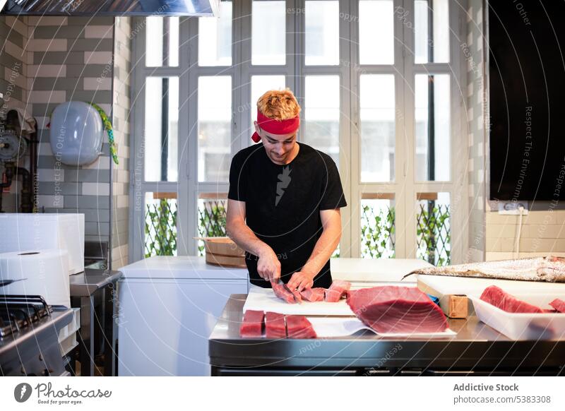 Focused young man standing and cutting tuna meat red fresh raw table concentrate focus kitchen male cook food fish culinary cuisine prepare glass inside